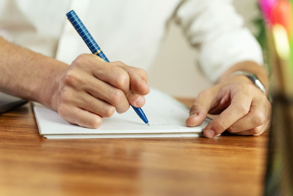 ノートにペンで数字やメモを書き込む男性の手元。ビジネスローンの借入計画や事業計画を練っている具体的な作業風景