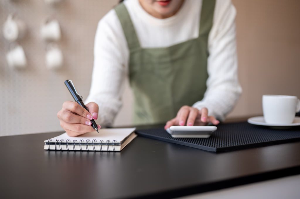 エプロン姿の女性店主がカウンターで電卓を叩きながらノートにメモを取る、店舗運営の資金繰りやビジネスローンの検討を想起させるシーン