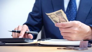 開業や事業拡大に必要な資金調達を検討する個人事業主が、政府系金融機関や民間からの融資を受けるために電卓でシミュレーションを行いながら現金を手にする様子