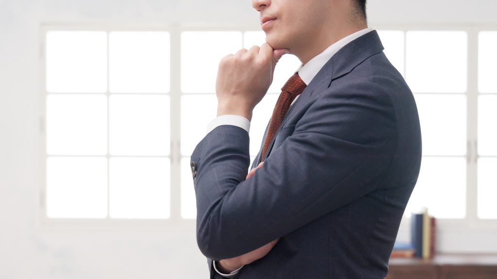 窓際で腕を組み、顎に手を当てて考え込むスーツ姿の男性。ローン審査なしで借入ができる方法を模索している様子をイメージ