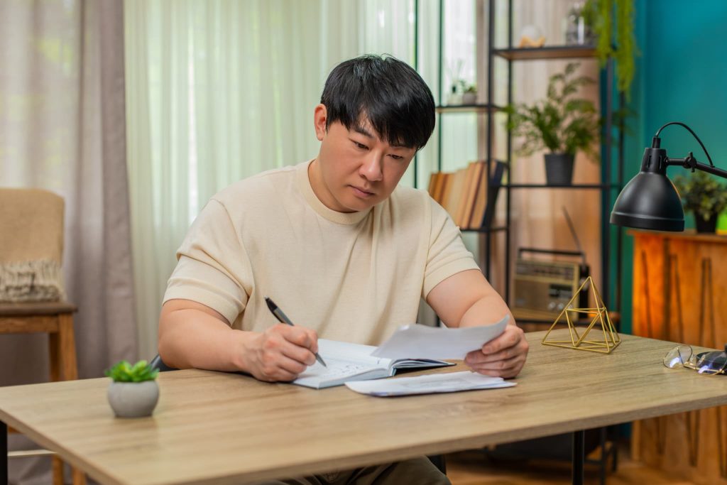 自宅のデスクで書類を真剣な表情で見つめ、ノートに記入する男性。個人事業主がビジネスローンの審査書類を準備する様子を描写