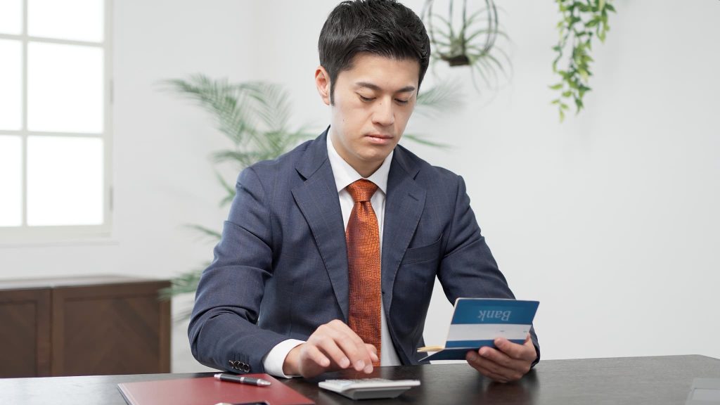 スーツ姿の男性がデスクで銀行の通帳を確認しながら電卓を叩く。法人向けのビジネスローン利用や口座管理をイメージさせる様子