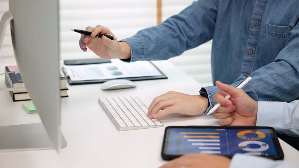 デスクトップPCの画面やタブレットのグラフを見ながら打ち合わせをするビジネスマン。個人事業主が売掛金の早期資金化について相談する様子