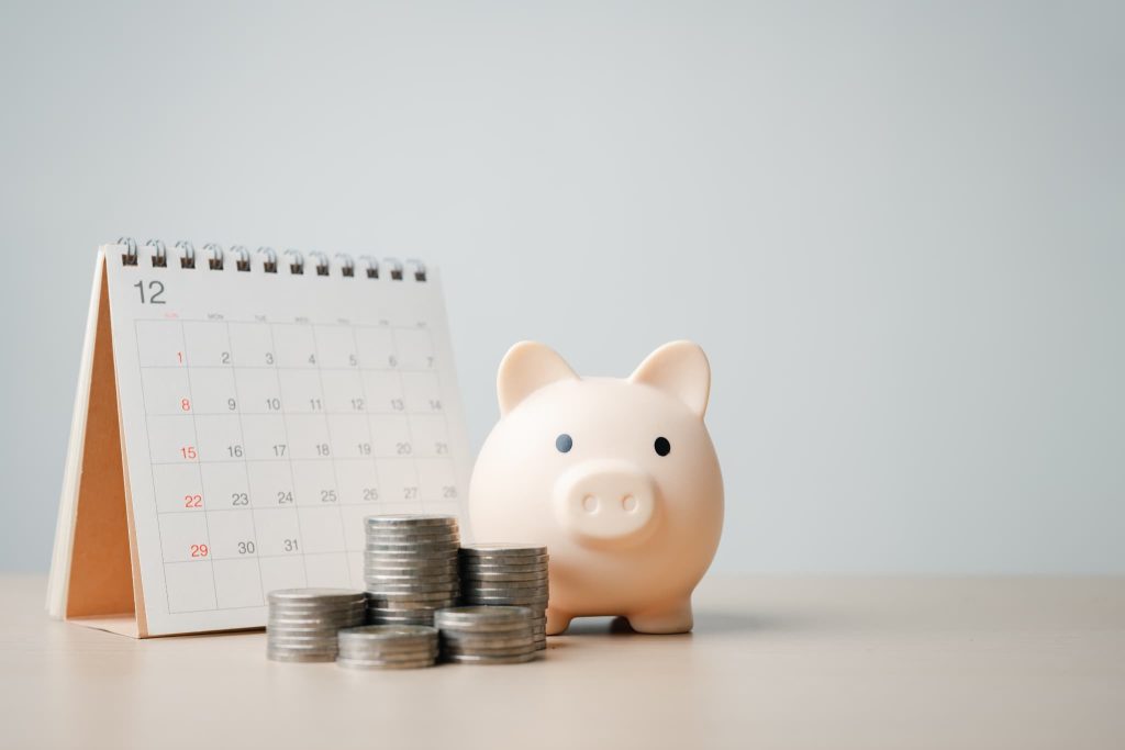 12月のカレンダーと積み上げられた硬貨の横に置かれた豚の貯金箱。毎月の支払いや資金繰りが苦しい状況を連想させるイメージ