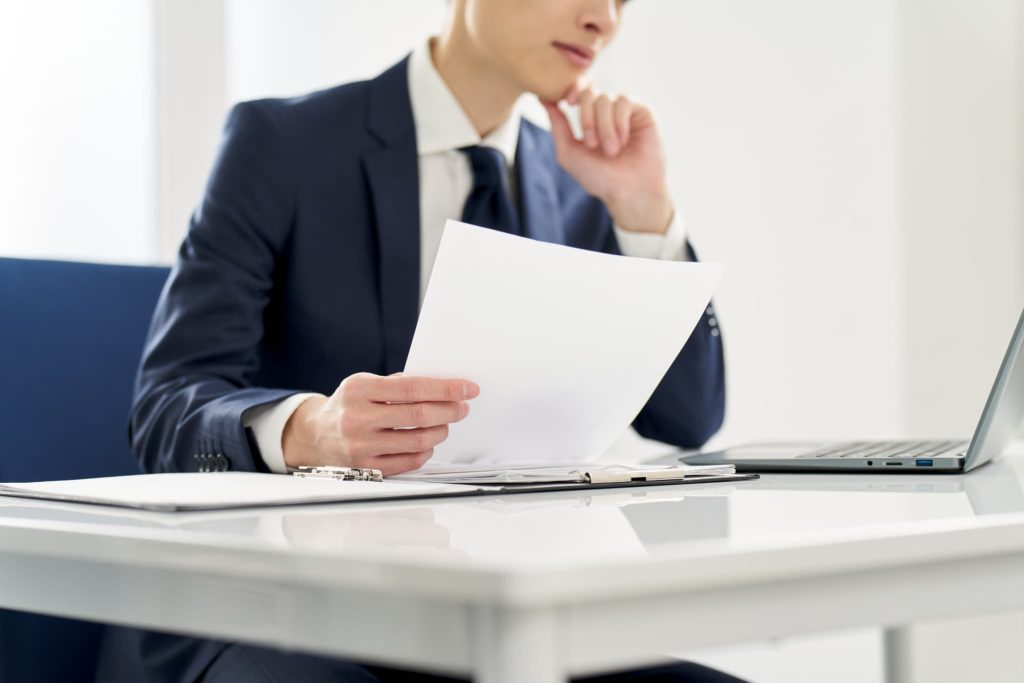 オフィスで書類を手に持ち内容を確認するスーツ姿の男性。少額ファクタリングの審査や必要書類の準備を進めているビジネスシーン