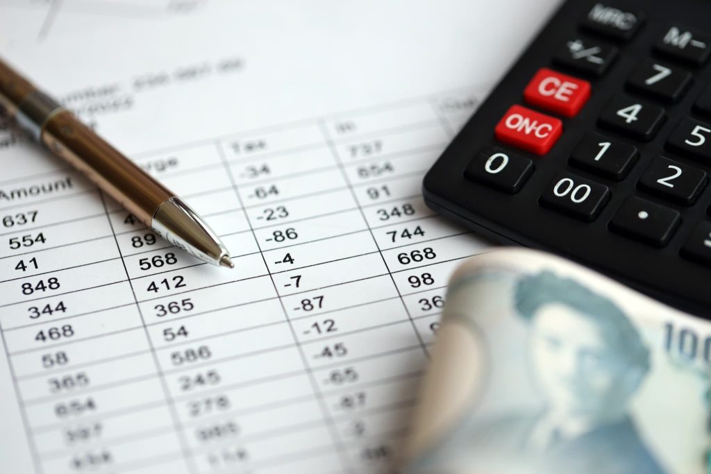 並んだ数字のリストの上に置かれたボールペンと電卓、そして一万円札のクローズアップ。個人事業主が資金調達を行うために必要な収支計画の策定や財務管理を表現