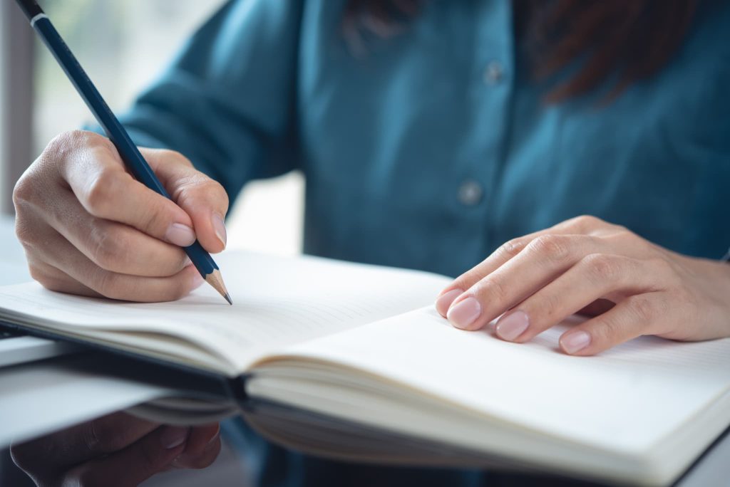 手元でノートに鉛筆で書き込みをする人物。運転資金の融資申し込みに向けた事業計画書の作成や、資金繰り表の整理を行っている様子