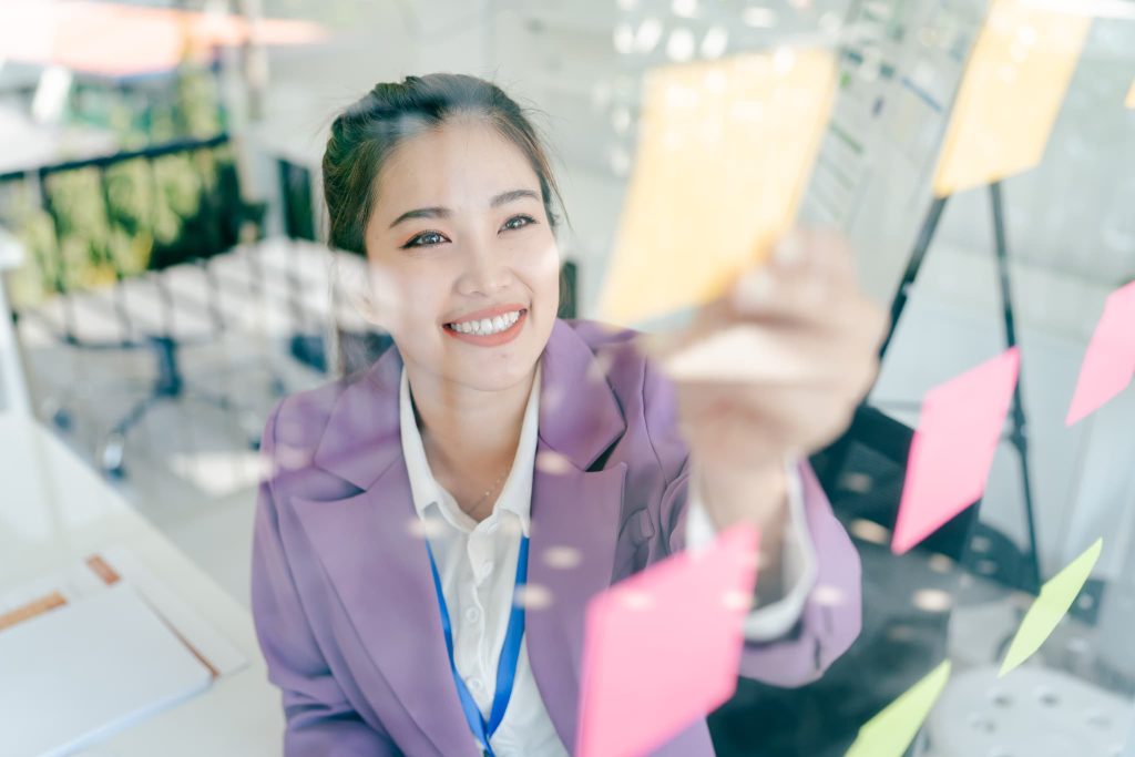 オフィスで付箋が貼られたガラスボードを前に笑顔を見せる女性起業家。個人事業主が融資を受けて事業拡大や新規プロジェクトに挑む明るい未来