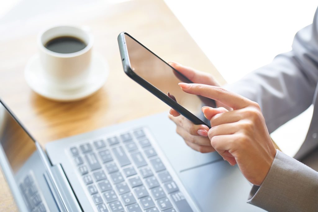 カフェでパソコンを開きながらスマートフォンを操作する女性。運転資金の融資についてオンラインで情報収集や申し込みを行っているビジネスパーソン