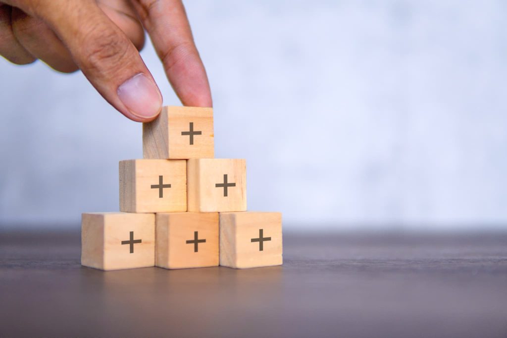 プラスの記号が印字された木製ブロックをピラミッド状に積み上げる手元。少額からのファクタリング利用による資金繰りの改善やメリットを表現