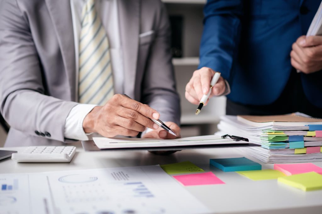 デスクに積まれた大量の書類を前に、二人のビジネスパーソンが内容を精査する様子。資金繰りとは複雑な経営判断を伴う実務であることをイメージ