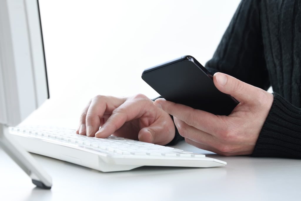 デスクトップパソコンのキーボードを打ちながらスマートフォンを操作する男性。オンラインで完結する少額ファクタリングの手続きや申し込み風景