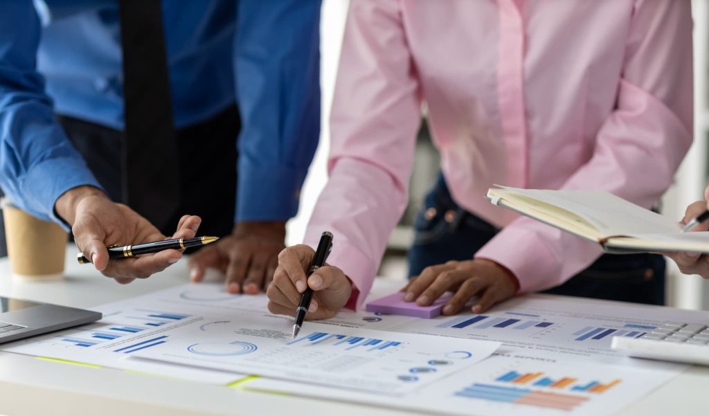 オフィスで複数の男女がグラフや表が記載された資料を指差し確認する様子。事業成長に向けた現状分析や資金繰りとは何かを議論しているシーン