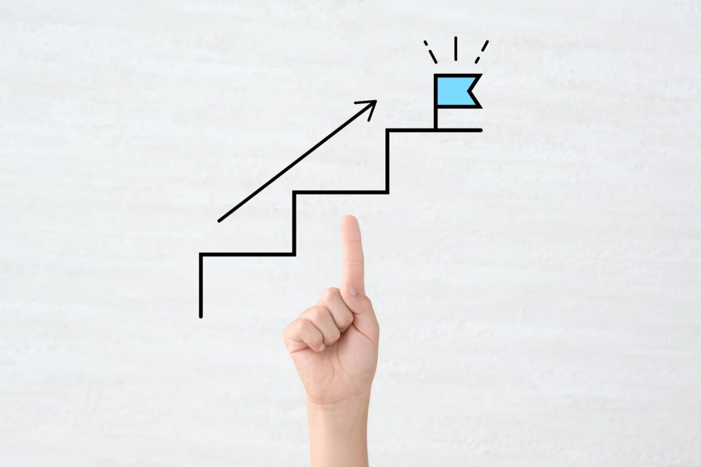 右肩上がりの階段とゴールを示す旗のイラストを指差す手。個人が資金調達を成功させ、事業を成長・ステップアップさせていく目標達成のプロセスを象徴