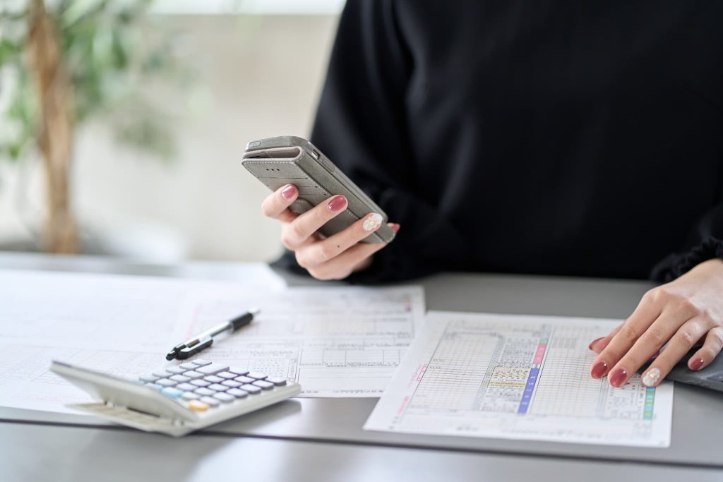 確定申告書や計算書類を前にスマートフォンを操作する女性。個人事業主が融資申し込みに必要な書類を揃え、スマホで手続きを進める様子