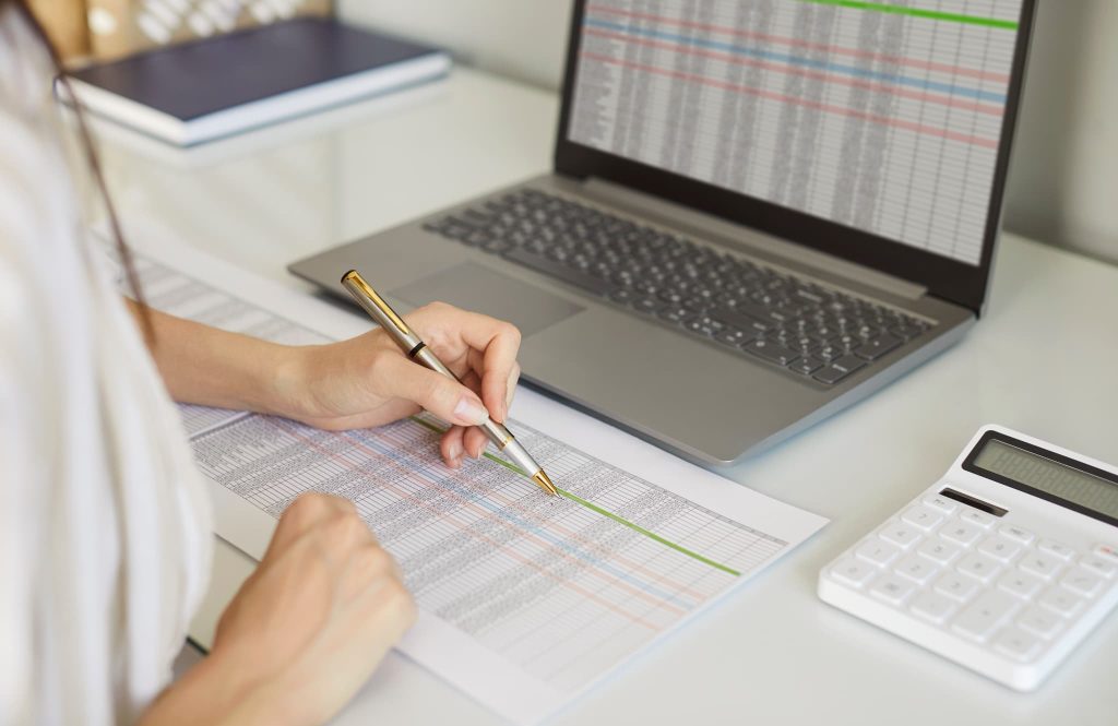 パソコンの横で詳細な数値が並ぶ表をペンでなぞりながら確認する手元。資金繰りとは正確な収支管理であること。を象徴する事務作業風景