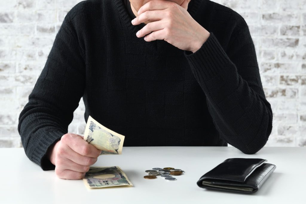 机の上の千円札と小銭を見つめ、顎に手を当てて悩む男性。個人事業主が融資を検討する際の資金繰りや手元の現金管理をイメージ