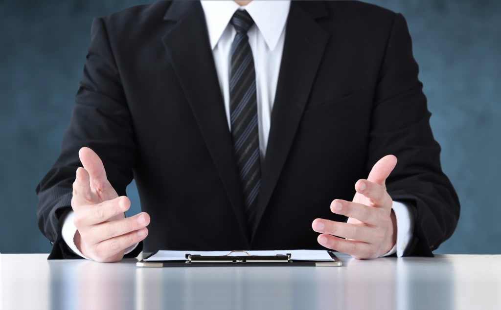 デスクで両手を広げて説明を行うスーツ姿の男性。ファクタリングによる資金調達の仕組みやメリットを専門家が分かりやすく解説しているビジネスシーン