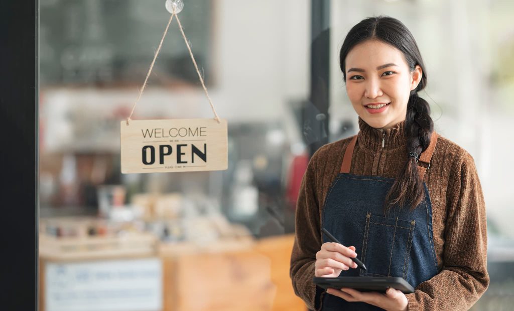 「OPEN」の看板が掛かったカフェの入り口で、タブレットを手に微笑む女性店主。個人で開業する際の資金調達や、店舗運営に必要な運転資金の準備をイメージ