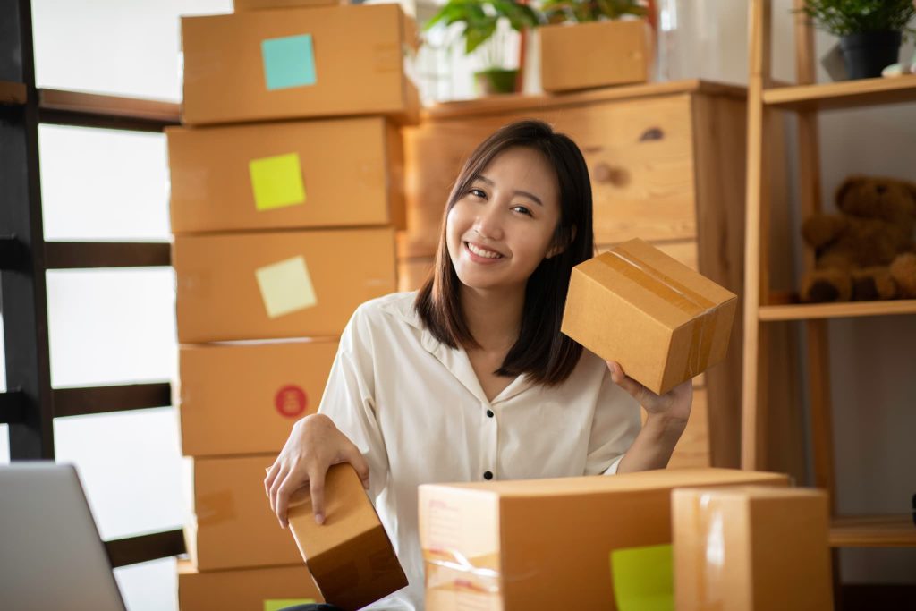 積み上げられた段ボールを背に笑顔を見せる女性。ネットショップなどの物販を営む個人事業主が、事業拡大や在庫確保のために運転資金の融資を検討するシーン