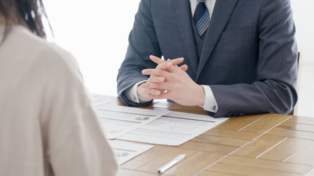 机を挟んで資料を前に話し合うスーツ姿の男性と相談者。個人事業主が融資担当者から運転資金の調達について専門的なコンサルティングや相談を受けている場面