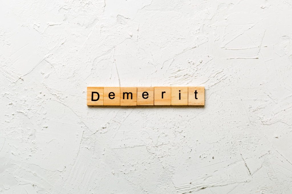 白い漆喰のような背景に置かれた「Demerit」の文字ブロック。法人ビジネスローンを契約する前に確認すべきデメリットや注意点を解説するシーンに最適