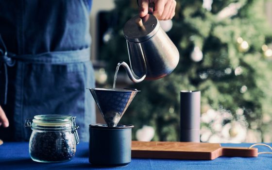 カフェの起業・開業手順を解説するイメージ写真。青いエプロンをつけたスタッフが、ポットからお湯を注ぎ、ハンドドリップでコーヒーを抽出している