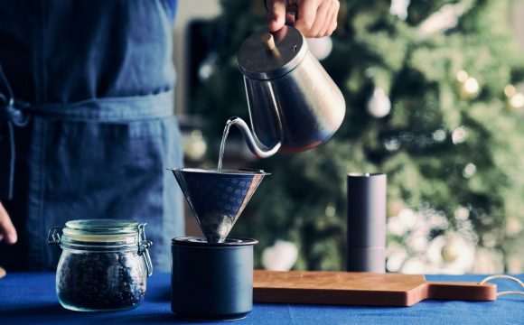 カフェの起業・開業手順を解説するイメージ写真。青いエプロンをつけたスタッフが、ポットからお湯を注ぎ、ハンドドリップでコーヒーを抽出している