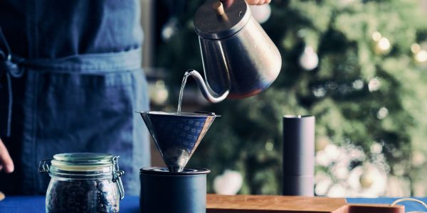 カフェの起業・開業手順を解説するイメージ写真。青いエプロンをつけたスタッフが、ポットからお湯を注ぎ、ハンドドリップでコーヒーを抽出している