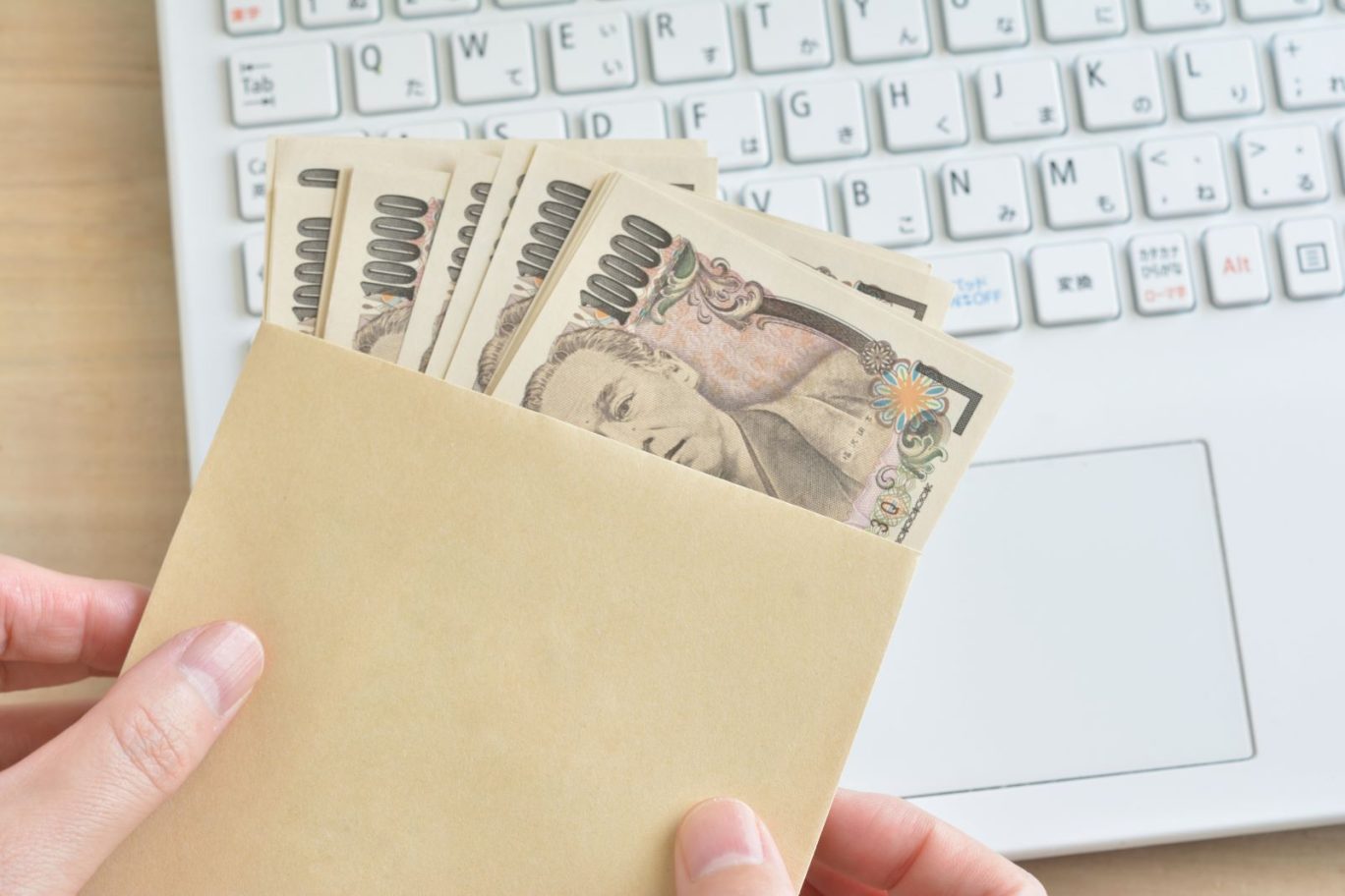 個人事業主が補助金として受け取る現金をイメージした写真。茶封筒に複数の1万円札が入っており、背景には作業中のノートパソコンが見えている