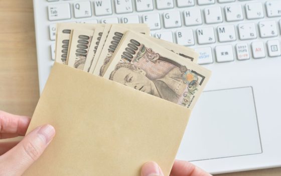 個人事業主が補助金として受け取る現金をイメージした写真。茶封筒に複数の1万円札が入っており、背景には作業中のノートパソコンが見えている
