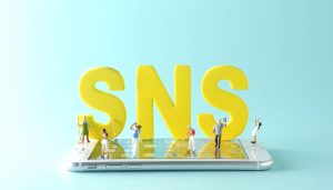 SNS広告とは何かを解説する記事のメイン画像。スマートフォンの上に置かれた「SNS」の文字の周りにミニチュアの人々が立っている