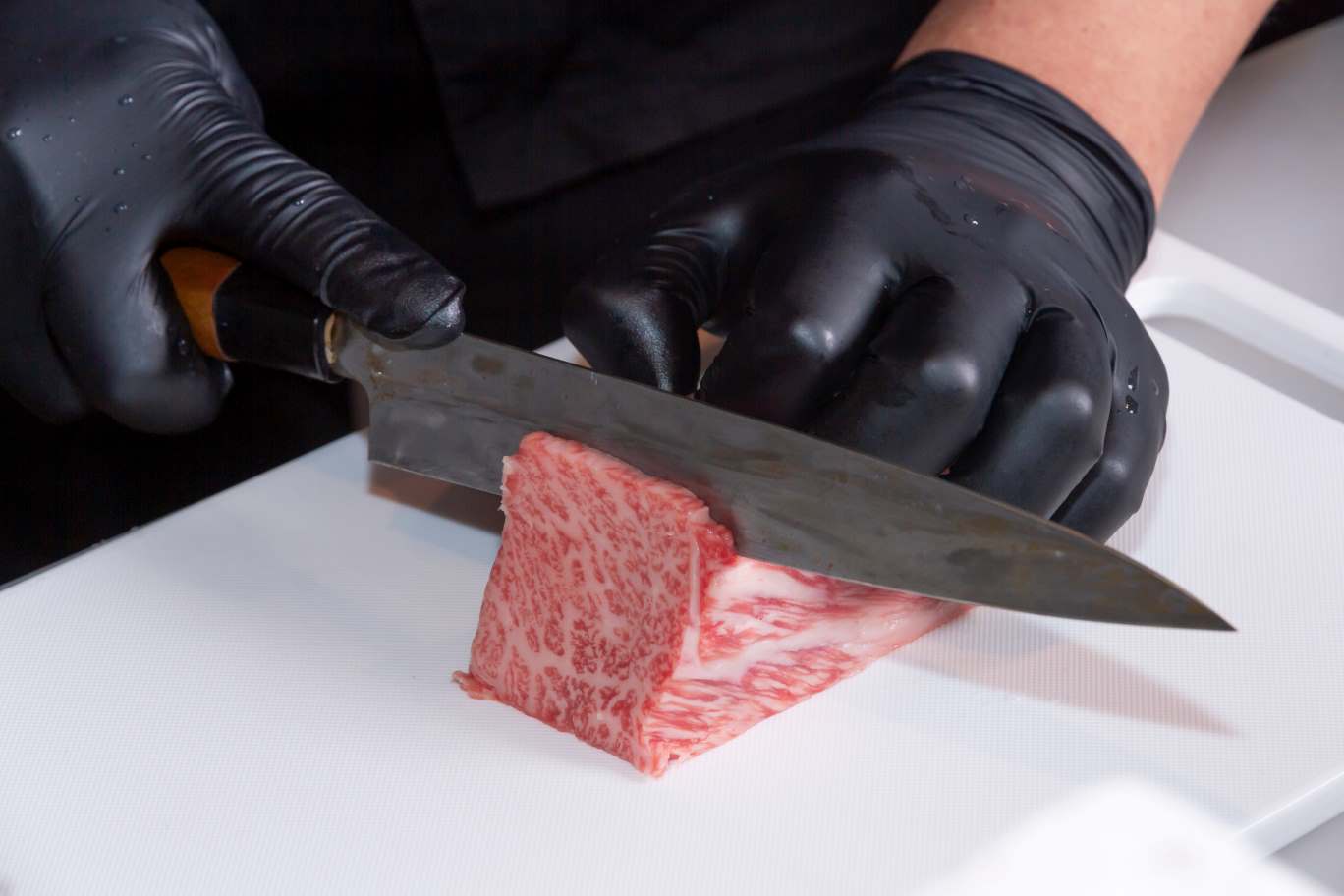 焼肉店経営の注意点を解説する写真。黒い手袋をしたスタッフが包丁で生肉をカットしている手元で、生肉を扱う上での厳格な衛生管理や品質管理の重要性を表現