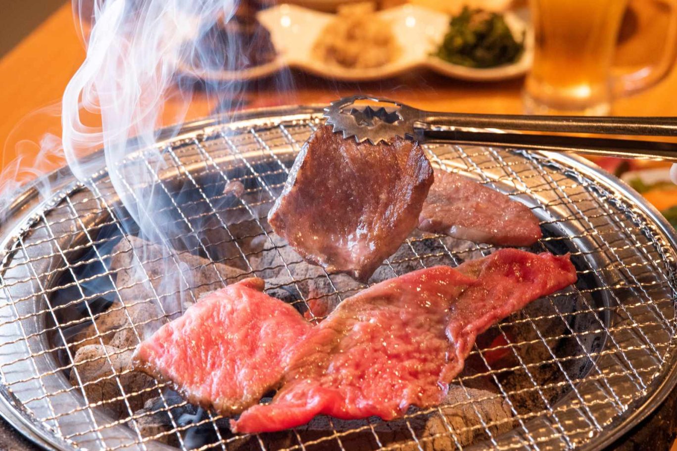 焼肉店の開業ノウハウを解説する記事のメイン画像。七輪の網の上でトングを使って赤身肉を焼いている、煙が立ち上る手元のアップ