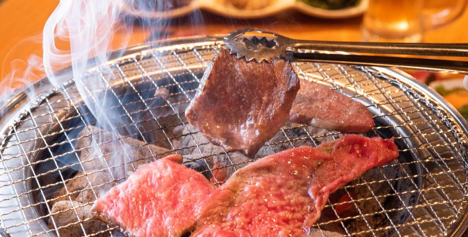 焼肉店の開業ノウハウを解説する記事のメイン画像。七輪の網の上でトングを使って赤身肉を焼いている、煙が立ち上る手元のアップ