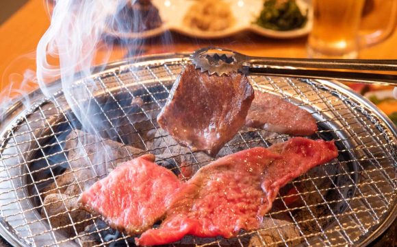 焼肉店の開業ノウハウを解説する記事のメイン画像。七輪の網の上でトングを使って赤身肉を焼いている、煙が立ち上る手元のアップ