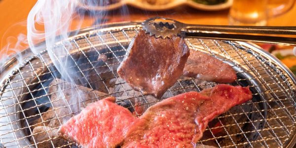焼肉店の開業ノウハウを解説する記事のメイン画像。七輪の網の上でトングを使って赤身肉を焼いている、煙が立ち上る手元のアップ