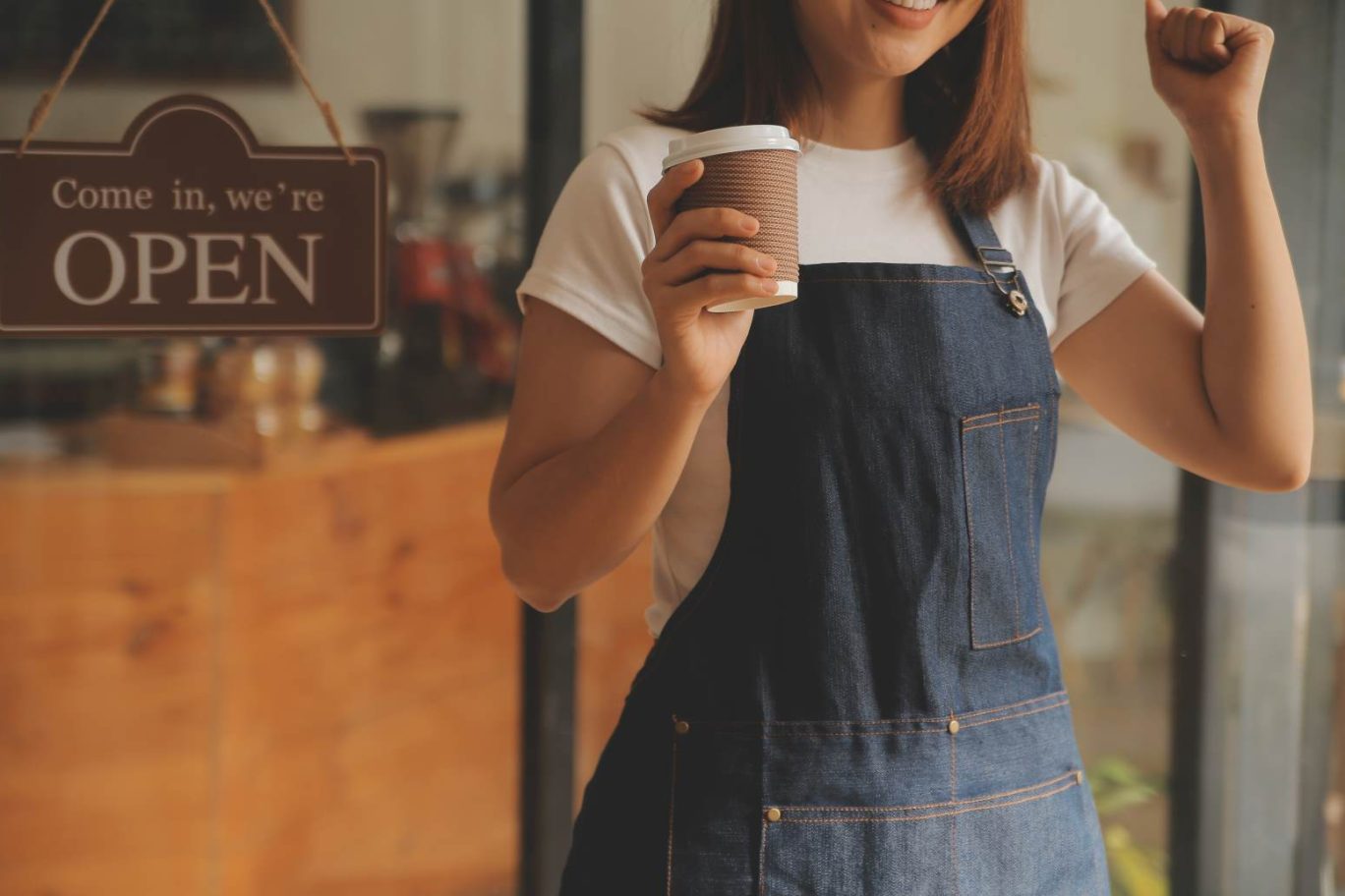 店舗開業をイメージした写真。エプロンをつけた女性が、「OPEN」と書かれた看板が掛かった店舗入口前で、コーヒーカップを片手にガッツポーズをしている