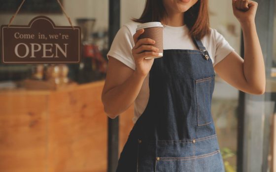 店舗開業をイメージした写真。エプロンをつけた女性が、「OPEN」と書かれた看板が掛かった店舗入口前で、コーヒーカップを片手にガッツポーズをしている