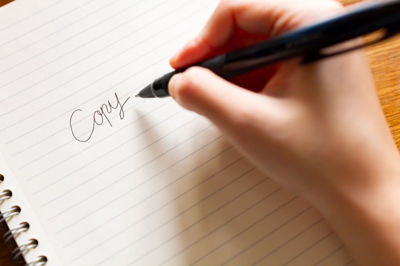 キャッチコピー制作をイメージした写真。白いノートに黒いペンで、「Copy」という文字を手書きしている様子