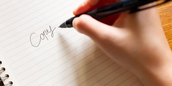 キャッチコピー制作をイメージした写真。白いノートに黒いペンで、「Copy」という文字を手書きしている様子