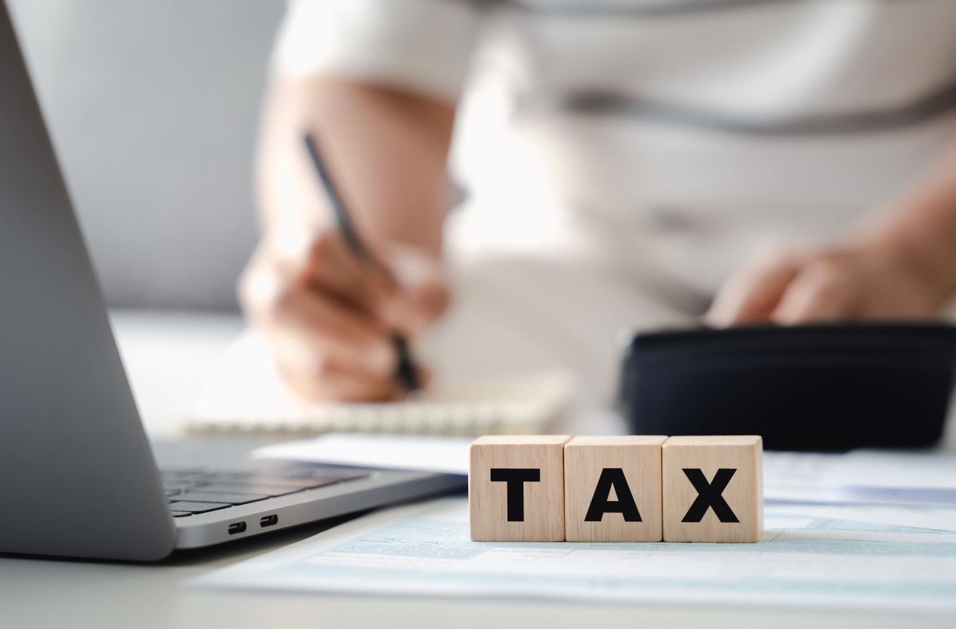 記事のメイン画像：手前には「TAX」の木製ブロックが置かれ、奥でパソコンや電卓を使って計算をしている人物がいる
