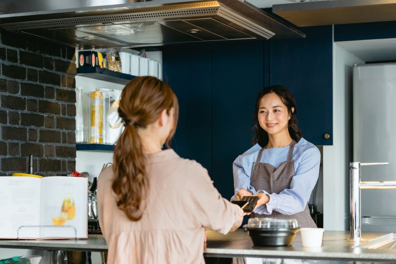 メイン画像：女性のアルバイトスタッフが飲食店のカウンターでキャッシュレス決済をしている