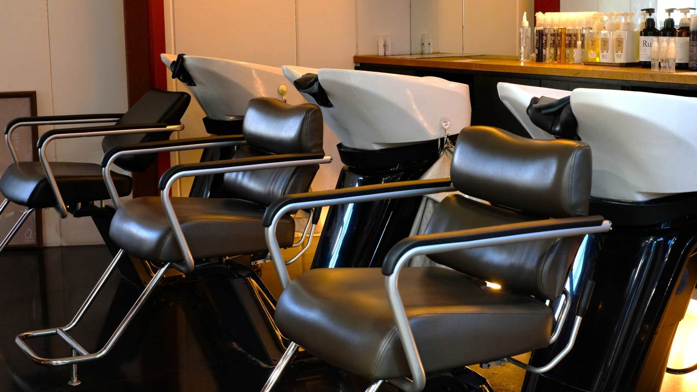 写真：美容室のシャンプースペース。黒いレザー調のシャンプーチェアと白いシャンプーボウルが3台連なって設置されており、奥のカウンターにはヘアケアボトルが並んでいる