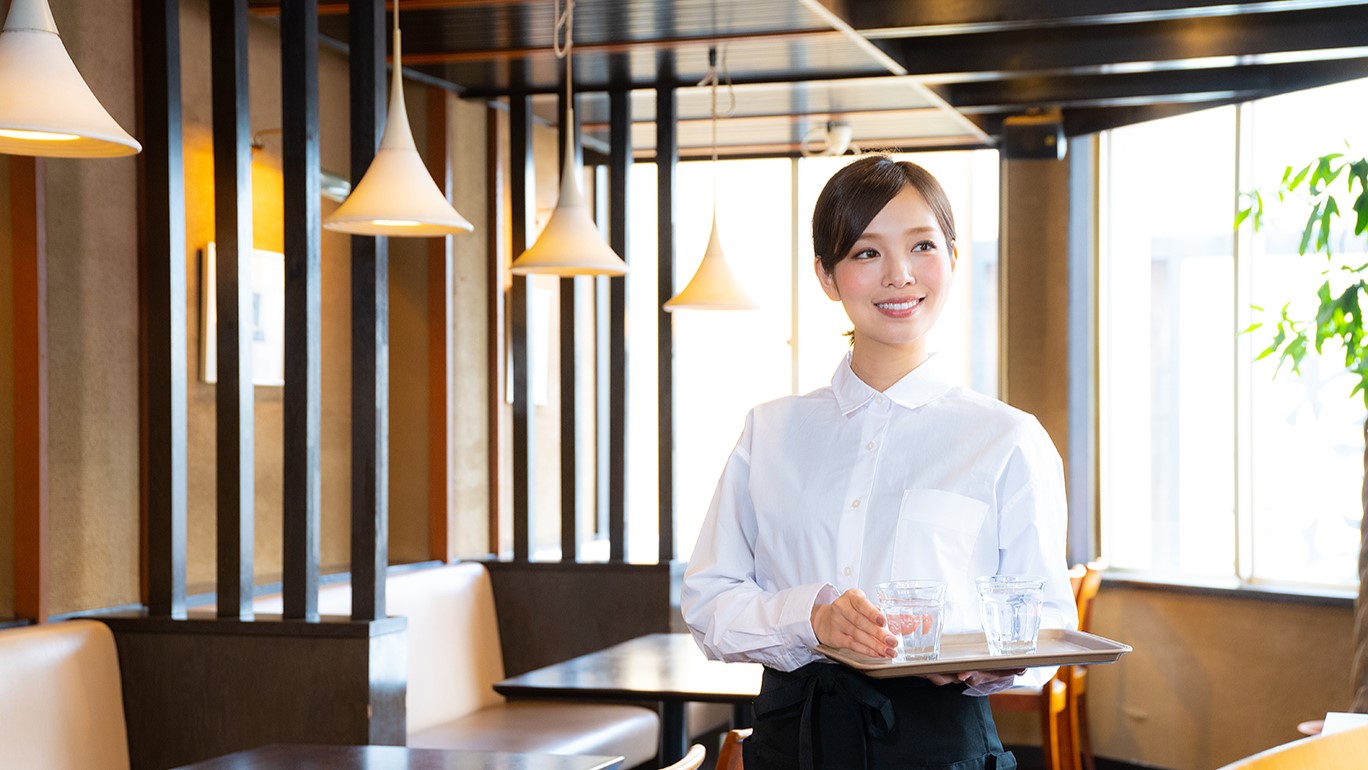 写真：お冷を乗せたトレイを持ち、店内で笑顔を見せる女性スタッフ