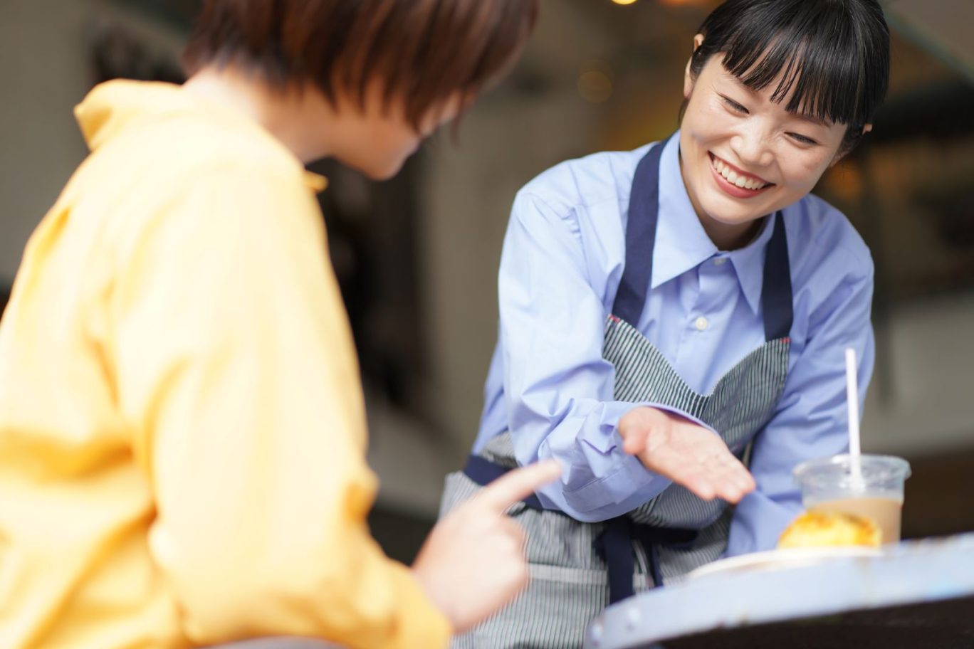 記事のメイン画像：カフェの店内にて満面の笑みでお客さまに商品を提供しているエプロン姿の女性スタッフ
